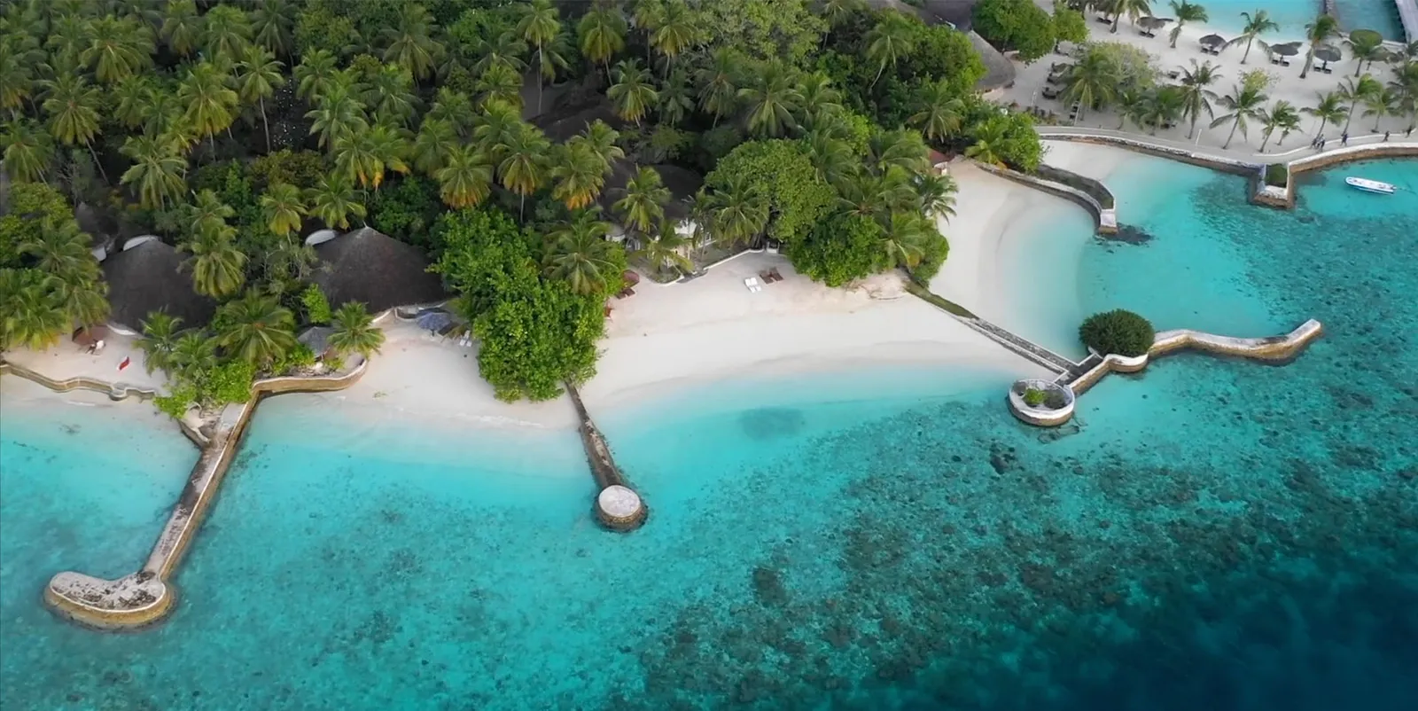 A stretch of shore from above: the white beach, the coral jetties and the reef showing through the lagoon.