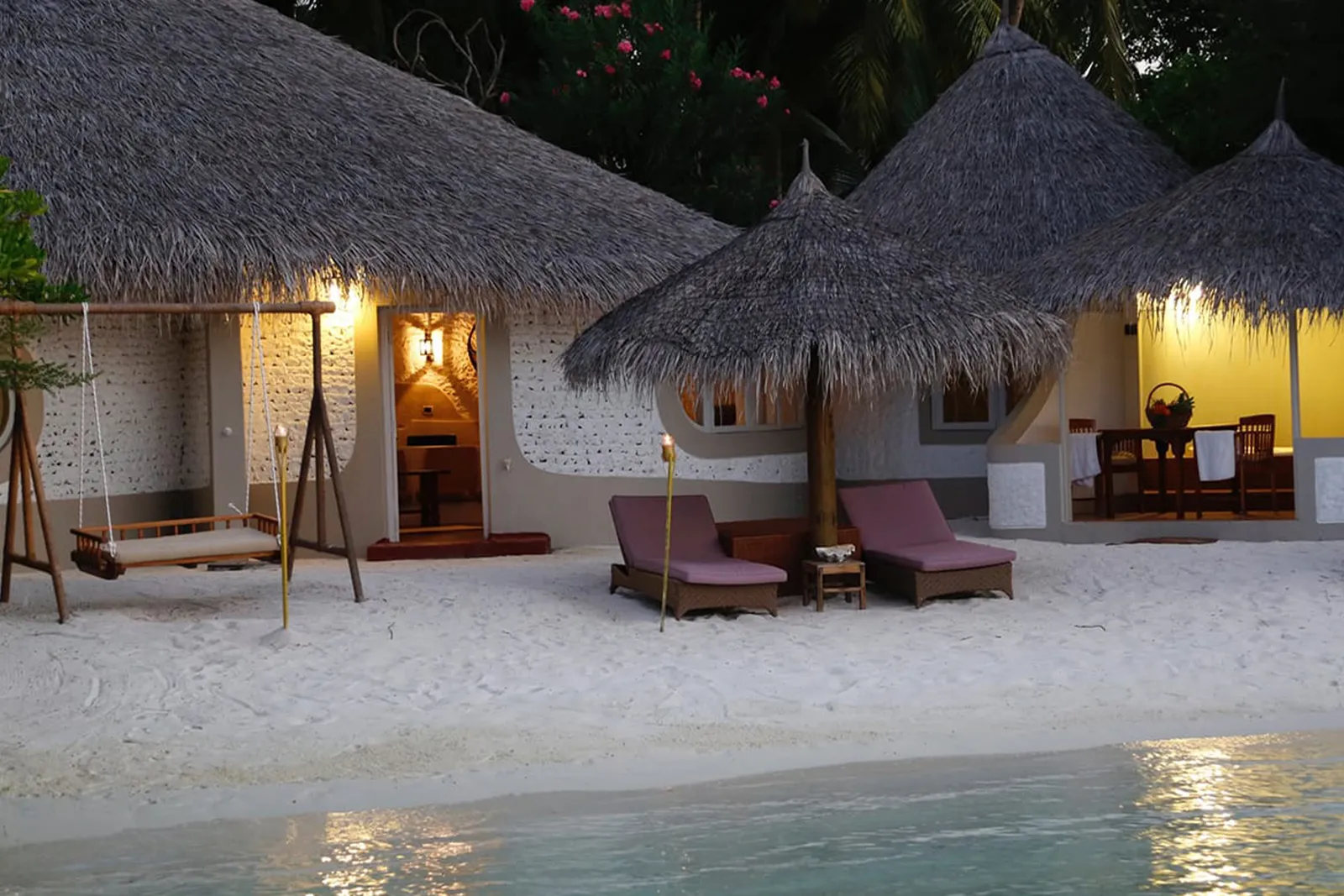 2 coral villas under thatch lit up at dusk, with a swing and loungers on the sand.
