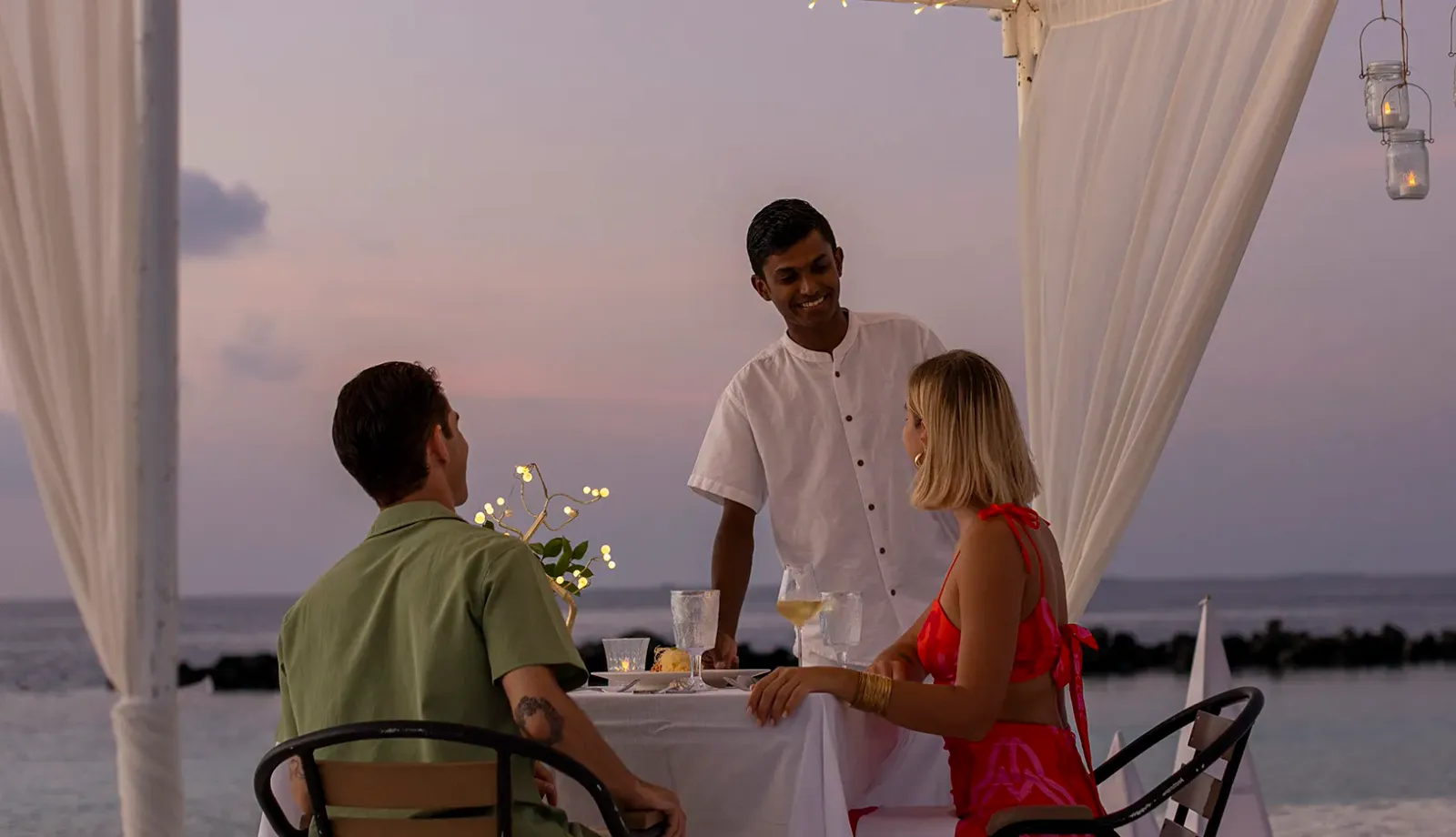 A beach dinner at sunset under a white-curtained canopy, a waiter at the table.