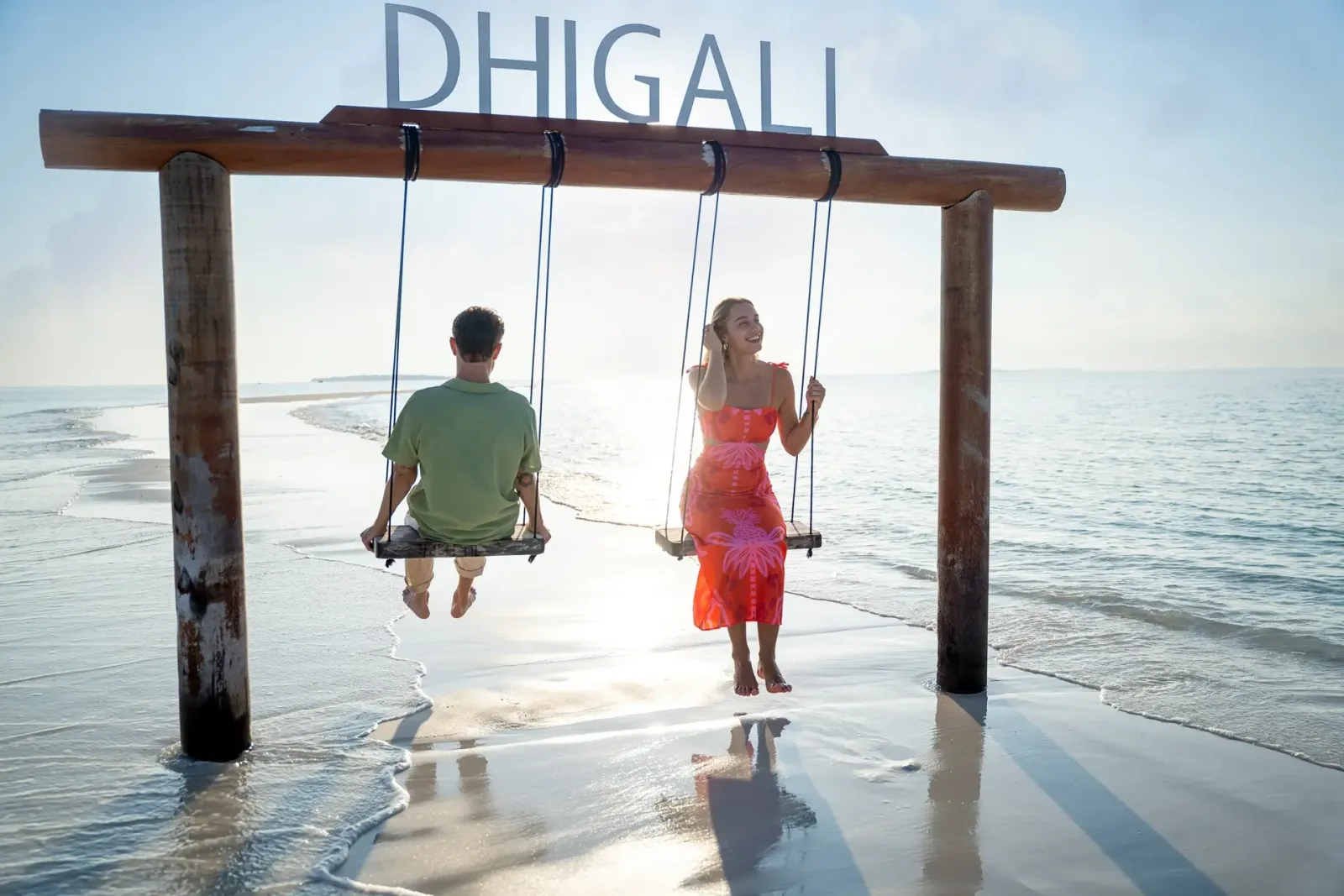 A couple on the swings at the water’s edge, under the DHIGALI sign, the sun low over the sea.