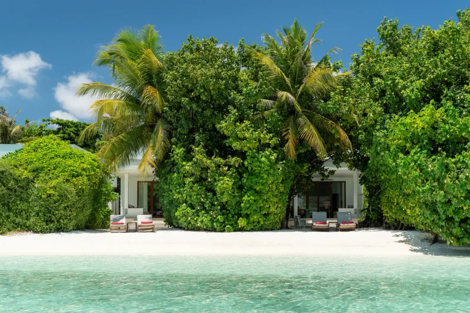 A beach villa hidden in the greenery, its loungers on the sand and the clear lagoon in front.