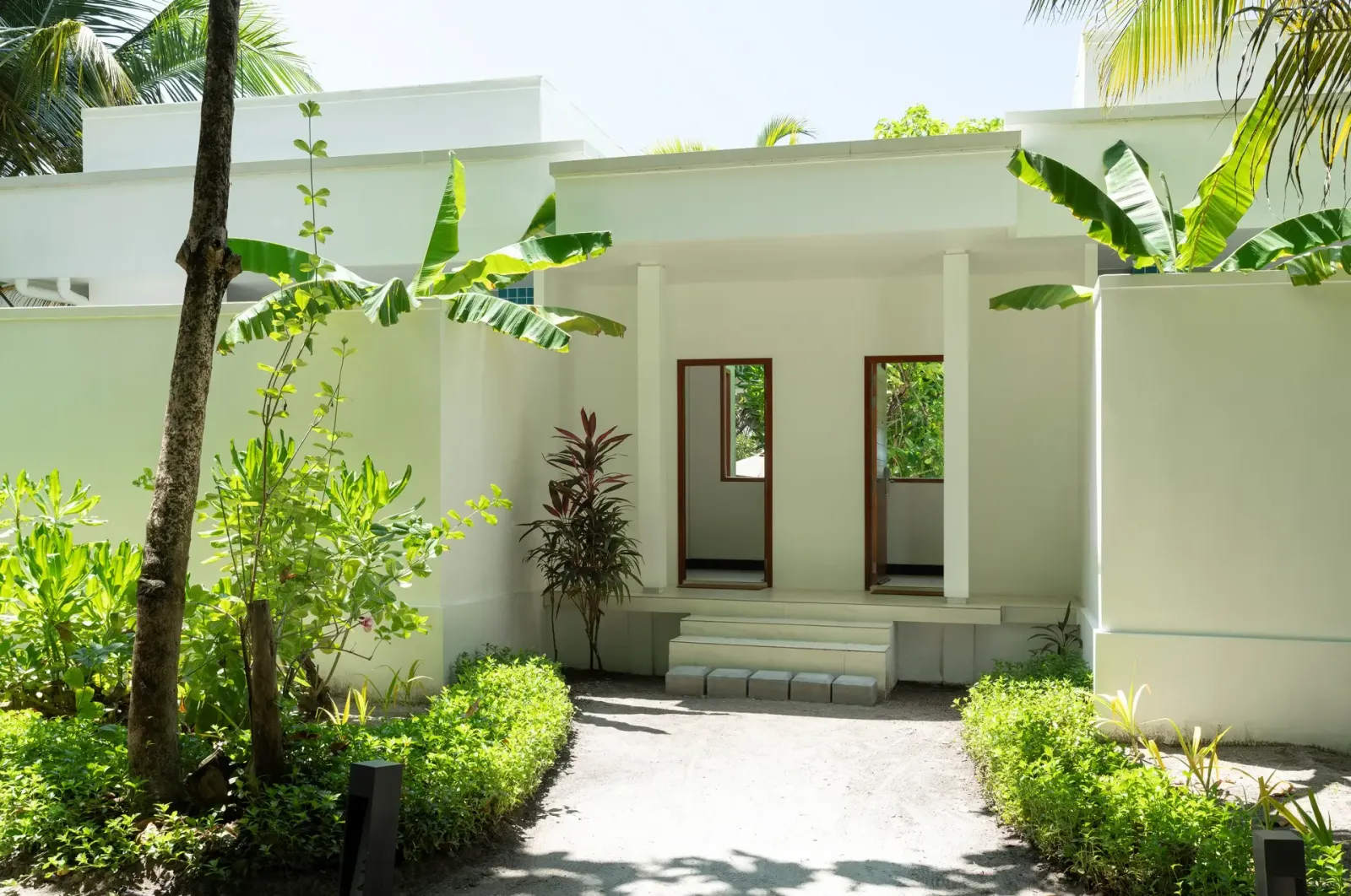 The entrance of a Two-Bedroom Deluxe Beach Bungalow, white, among banana plants and hedges.