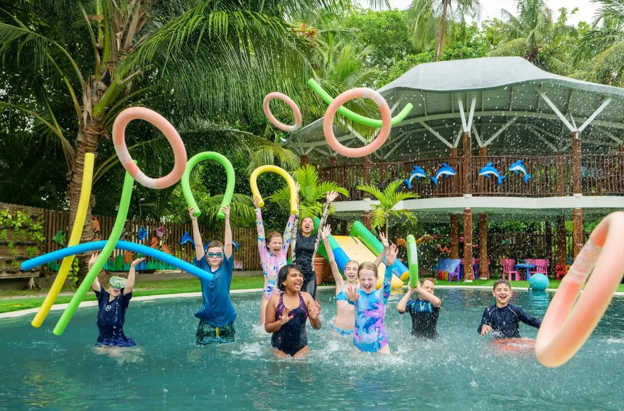 Children leaping with coloured pool noodles in Bageecha’s pool, under the palms