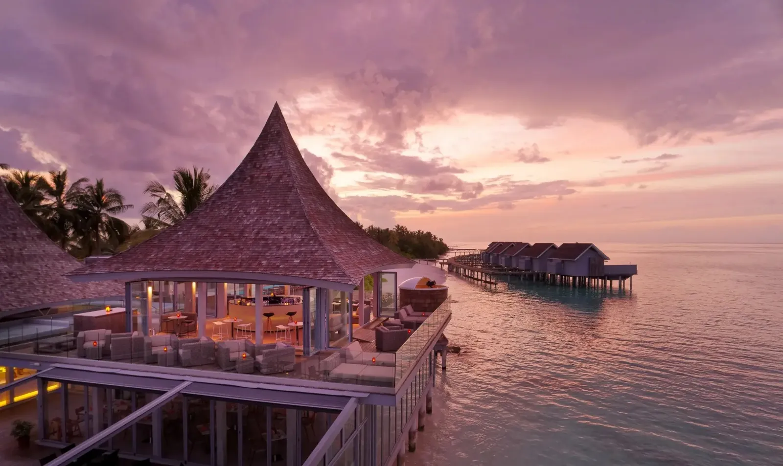 An overwater restaurant with a pointed roof, its lights on at dusk and the water villas behind