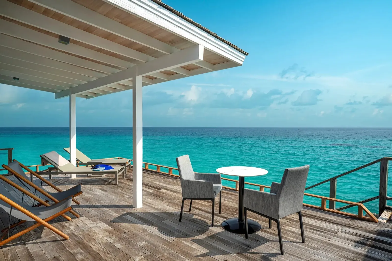 A Deluxe Water Villa’s deck over the sea, with loungers, a table for 2 and the turquoise ocean