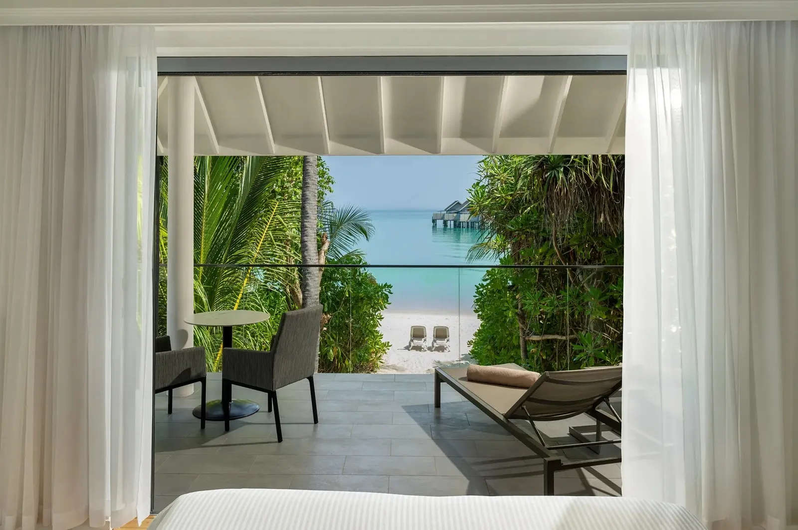 From a bedroom of the Two Bedroom Beach House: the veranda with 2 chairs and the beach through the greenery