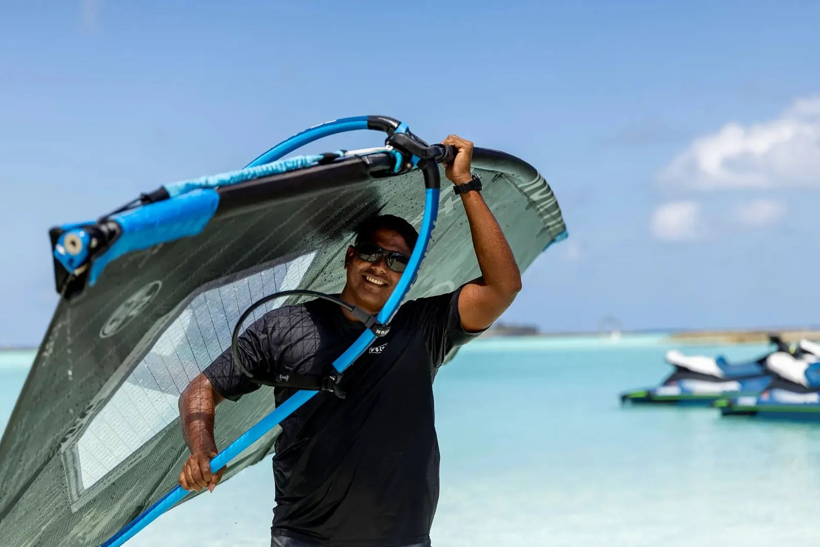 An instructor carries a windsurf board on the beach, jet skis behind