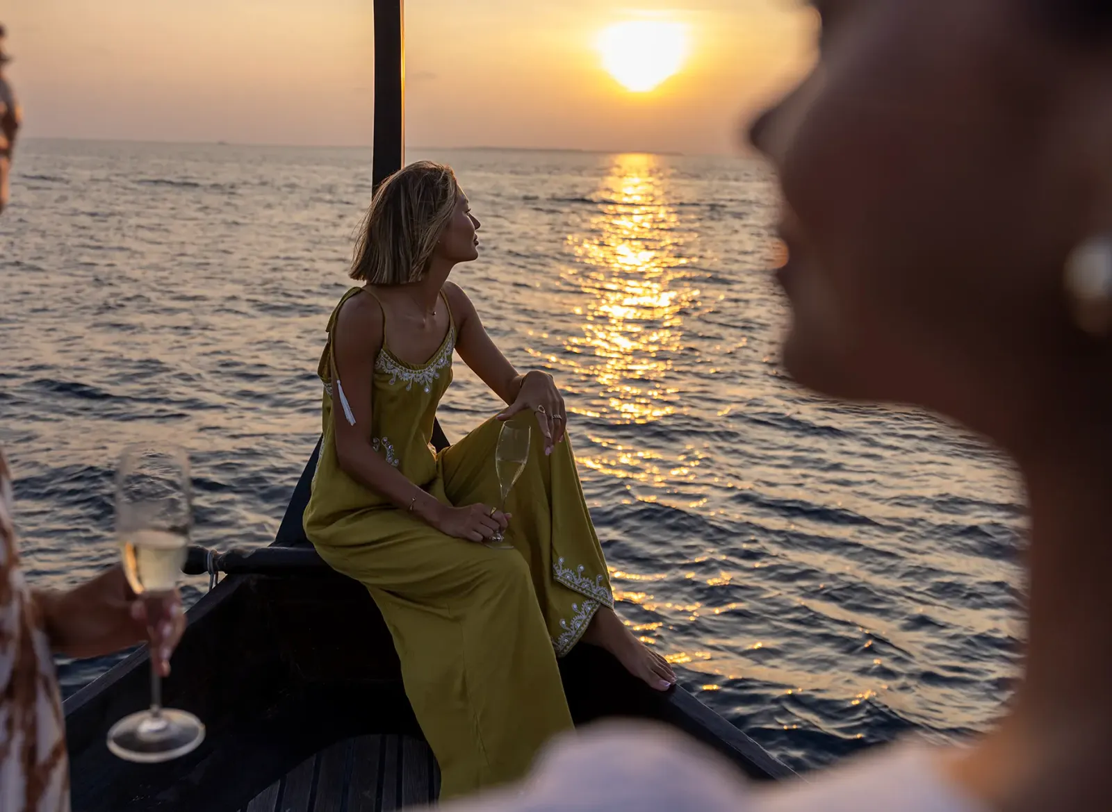 Un’ospite seduta a prua di un dhoni al tramonto, con un calice in mano.