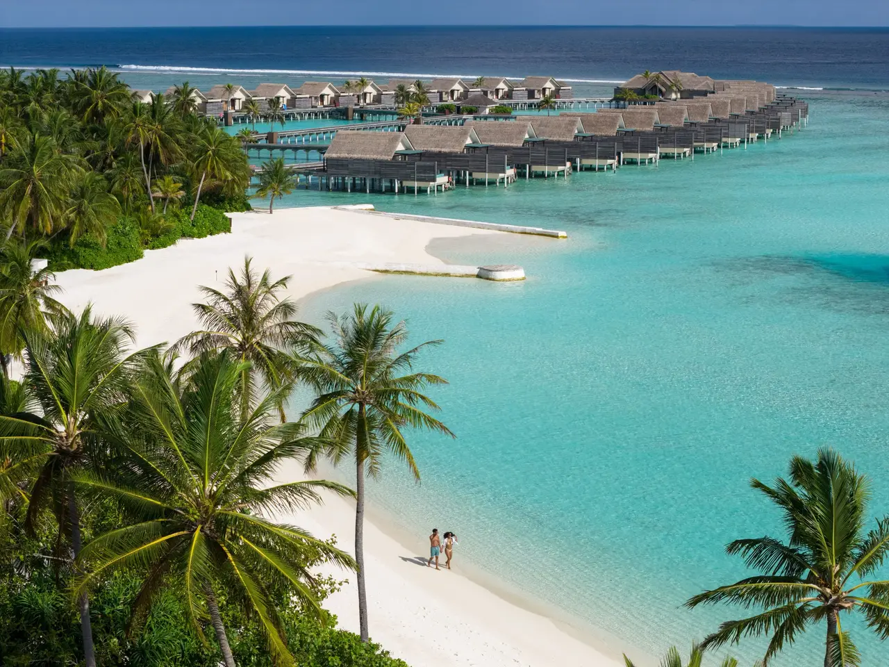 A couple walking on the beach of a cove, with the row of overwater villas behind