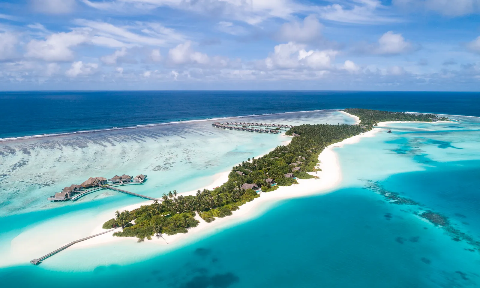 The long green island from above, with its white beach, the reef and the overwater pavilions at the end