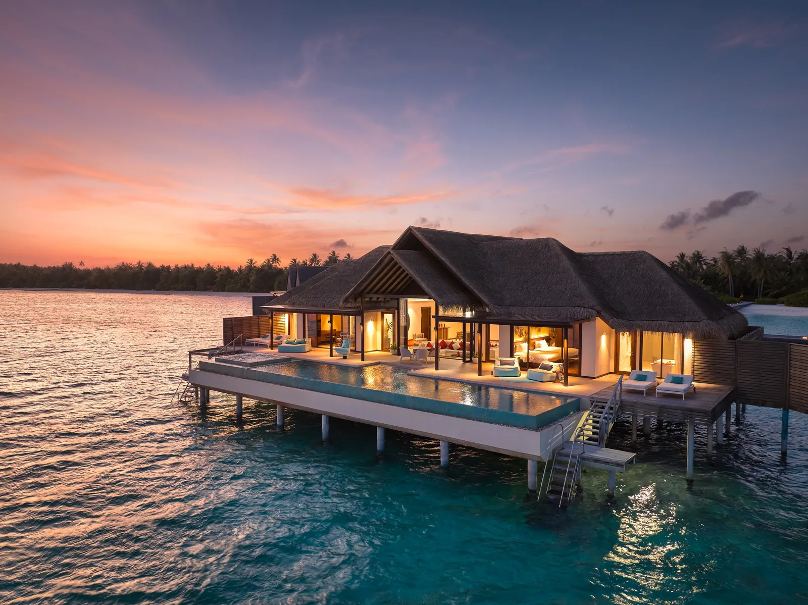 A 2-bedroom overwater pavilion at dusk, lights on and its pool lit