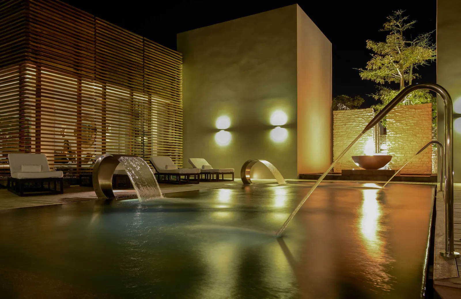 An outdoor pool at NIZUC Spa at night, with cascading water jets, white loungers and slatted timber walls.