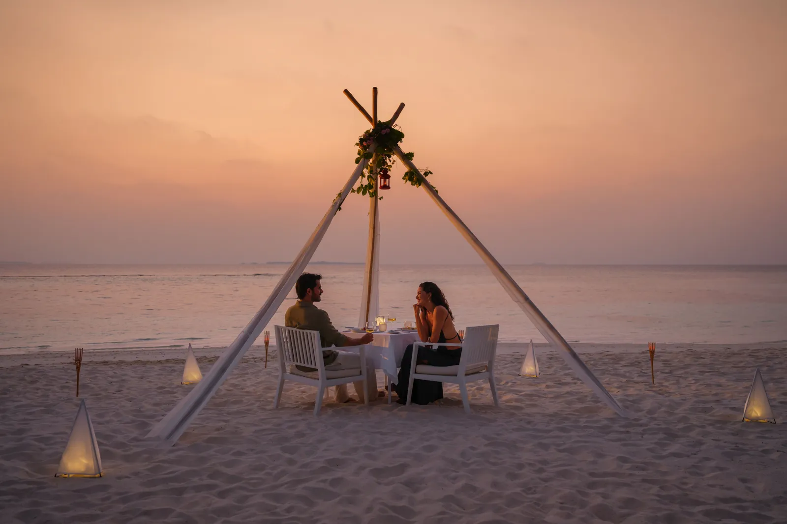 Dinner for 2 on the beach at sunset, under a wooden tripod hung with flowers, among lanterns on the sand.