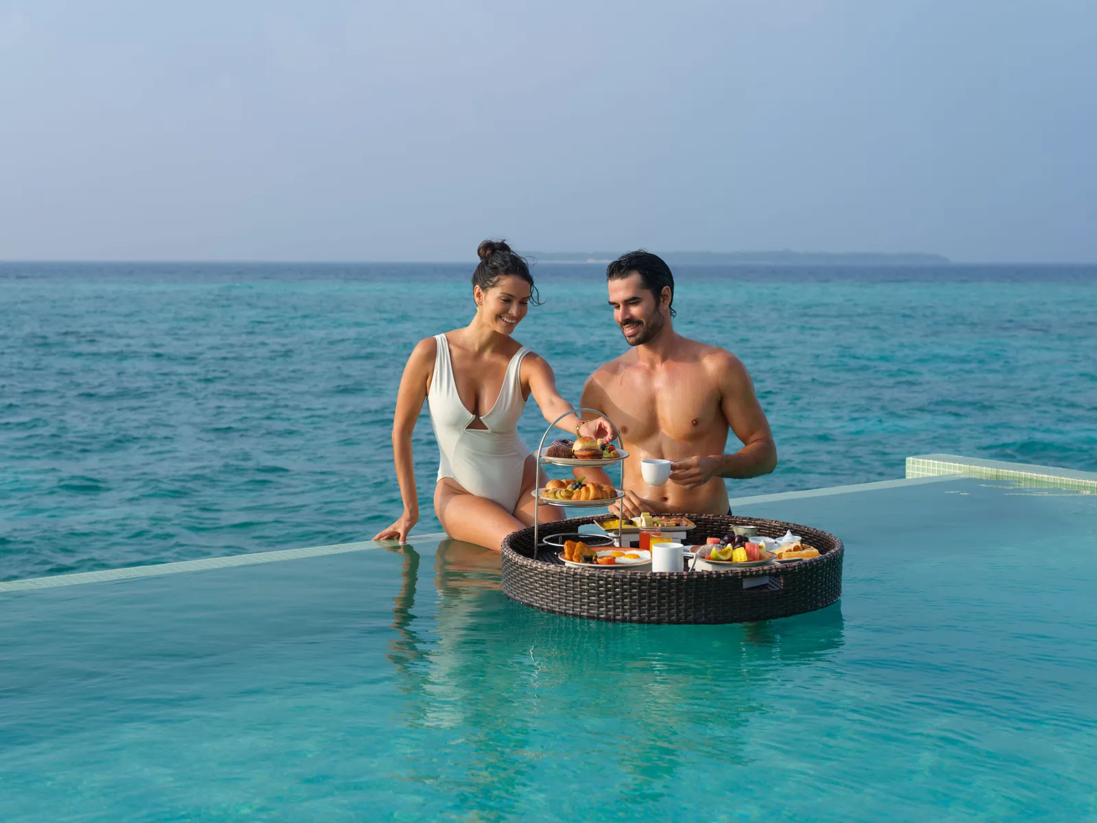 A couple breakfast in a villa pool from a floating tray, with the sea beyond the edge.