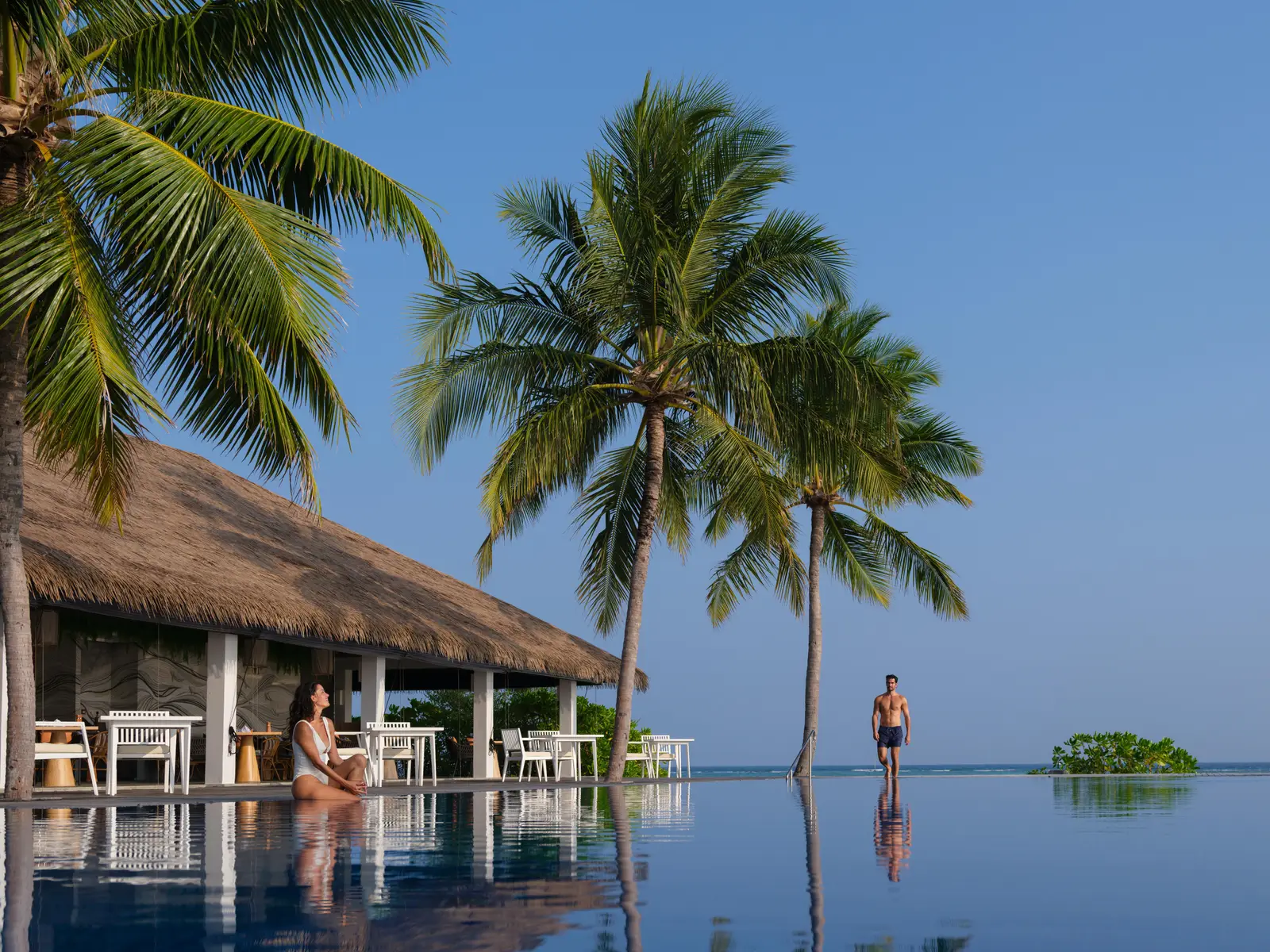 The infinity pool beside Thari: a woman seated on the edge, a man standing in the water and 3 palms reflected.