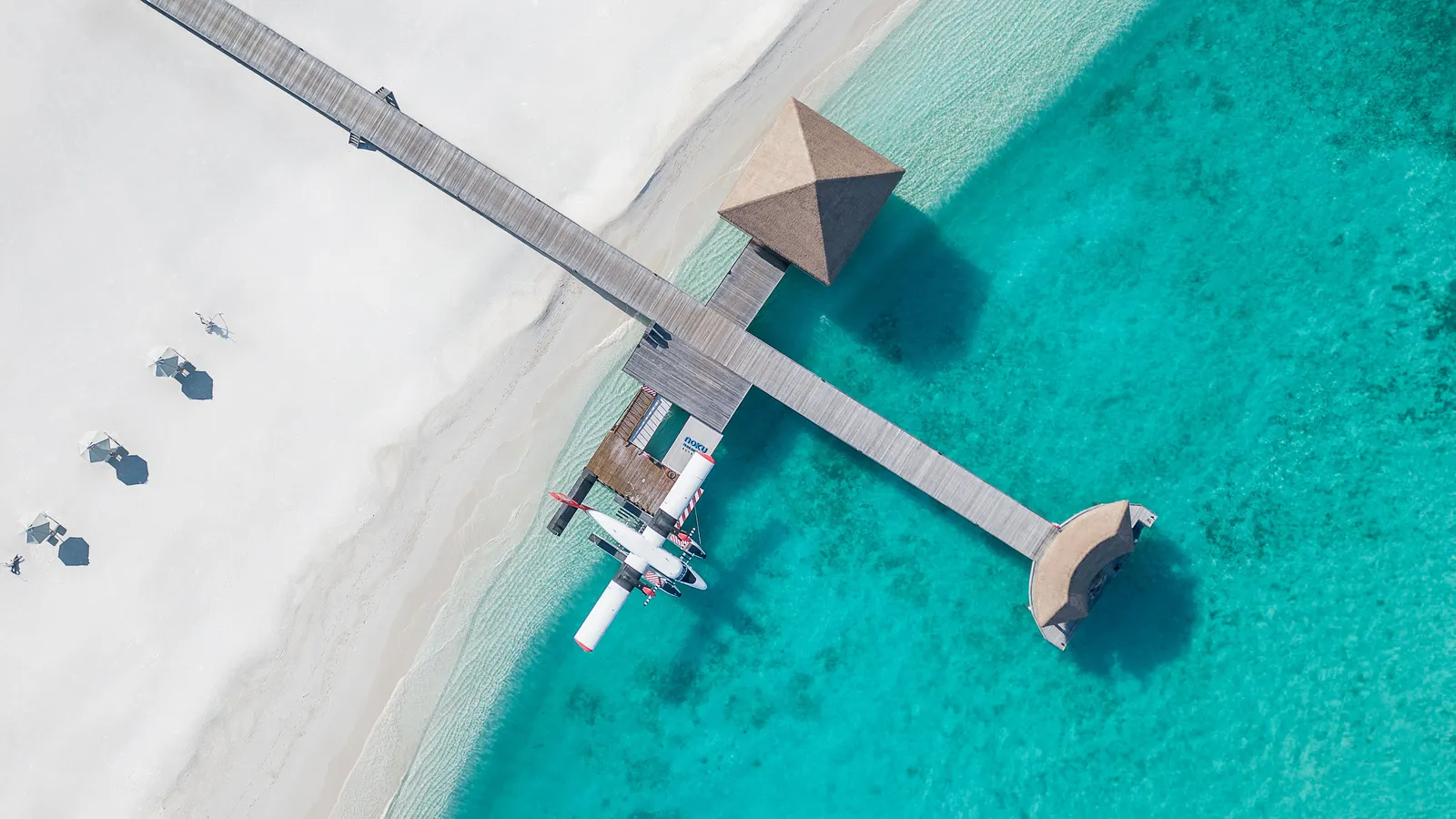 The jetty from above, with a seaplane moored at its platform, the white sand and the turquoise water.