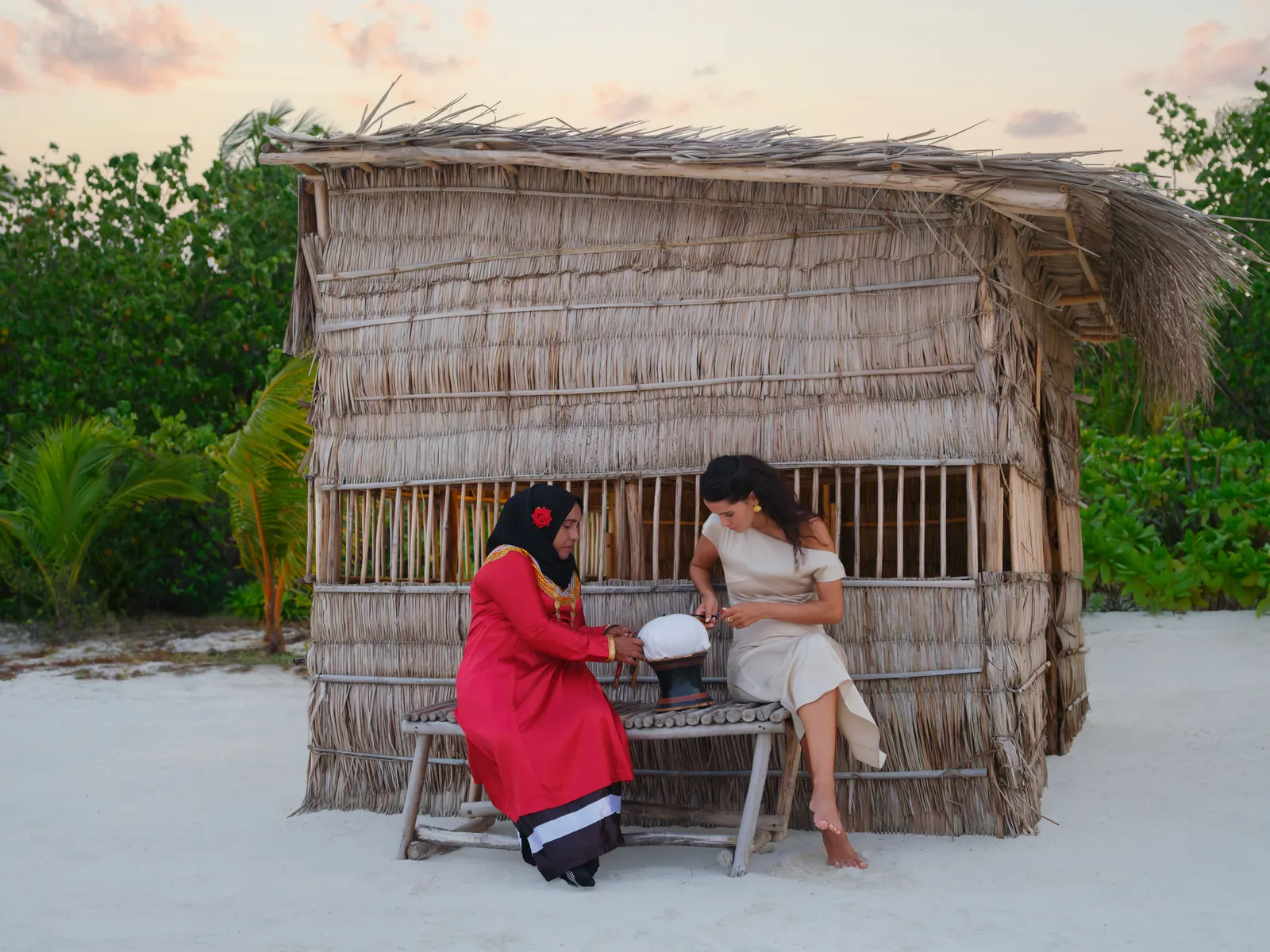 The Maldivian House: a palm-leaf hut on the sand, where a woman in traditional dress shows an object to a guest.
