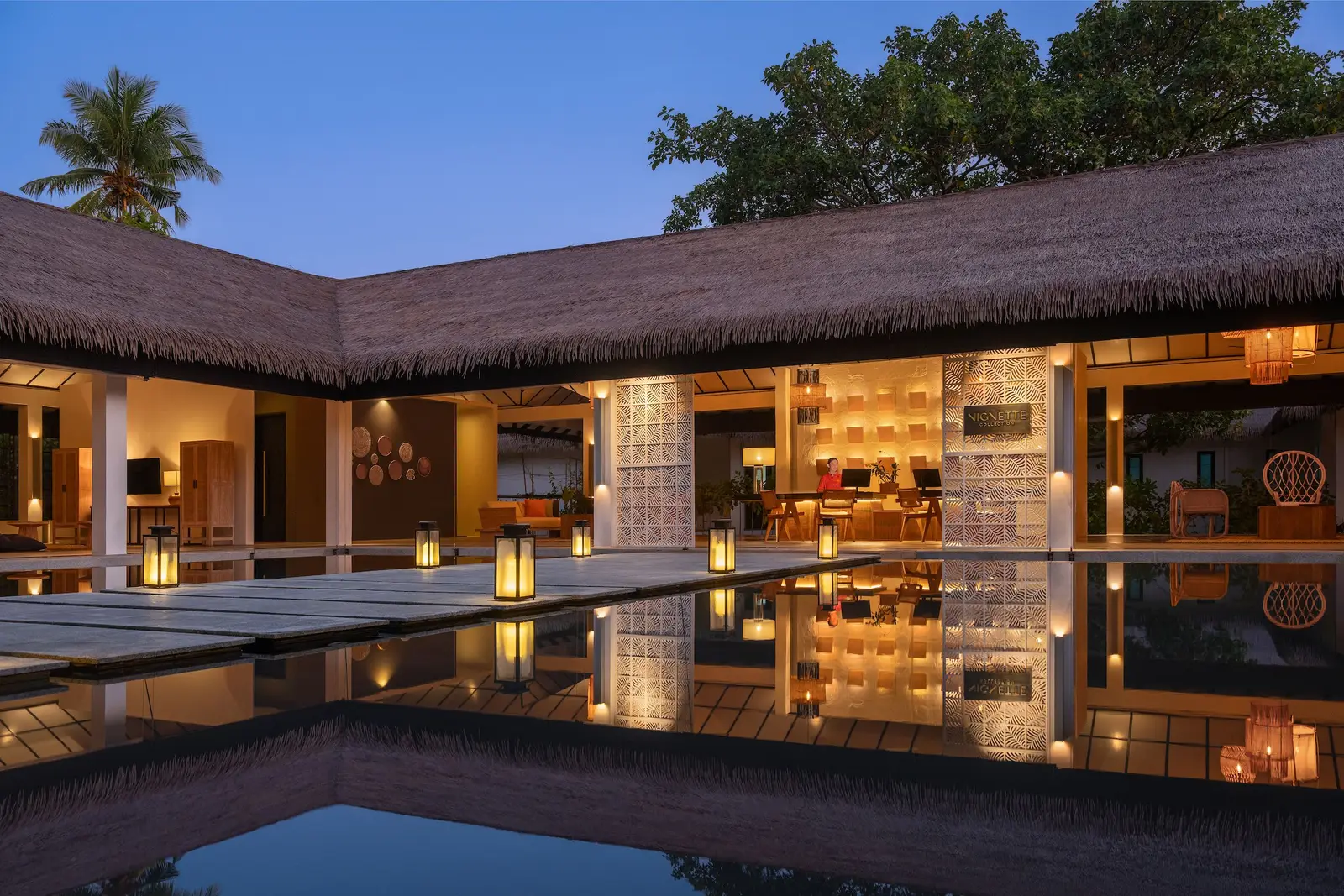 Reception at dusk, under its thatched roof, with the lanterns lit and the pool of water in front.