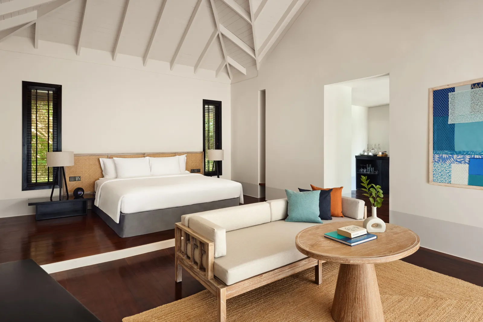 A beach villa bedroom: the king bed, the sofa, the round table and the high white timber ceiling.
