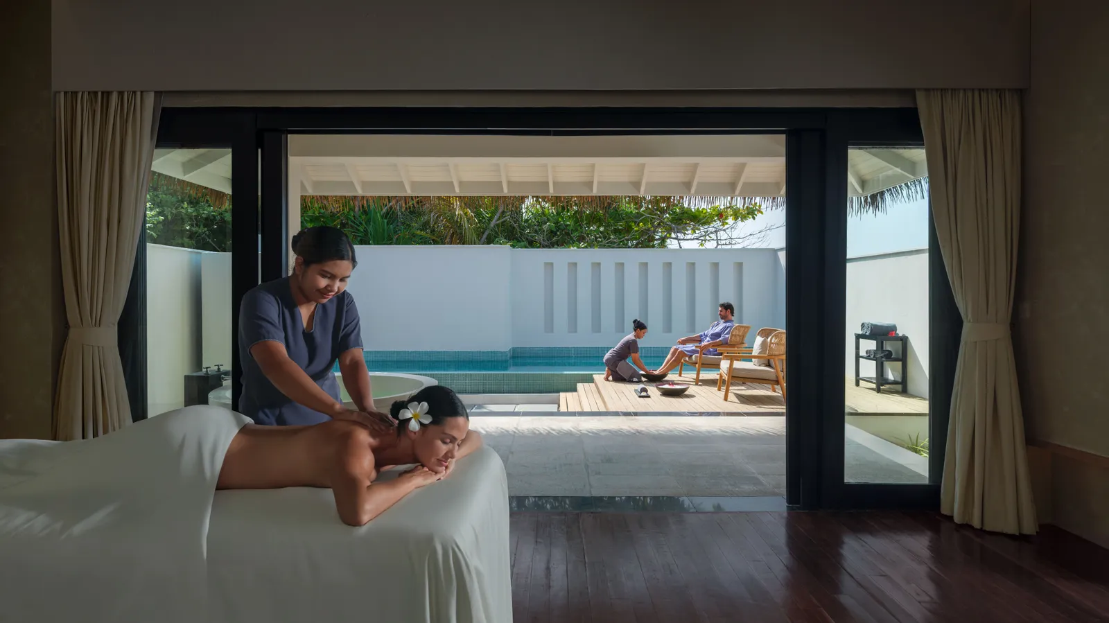 A massage in a spa room, the glass doors open onto a small pool and a patio where 2 people rest.