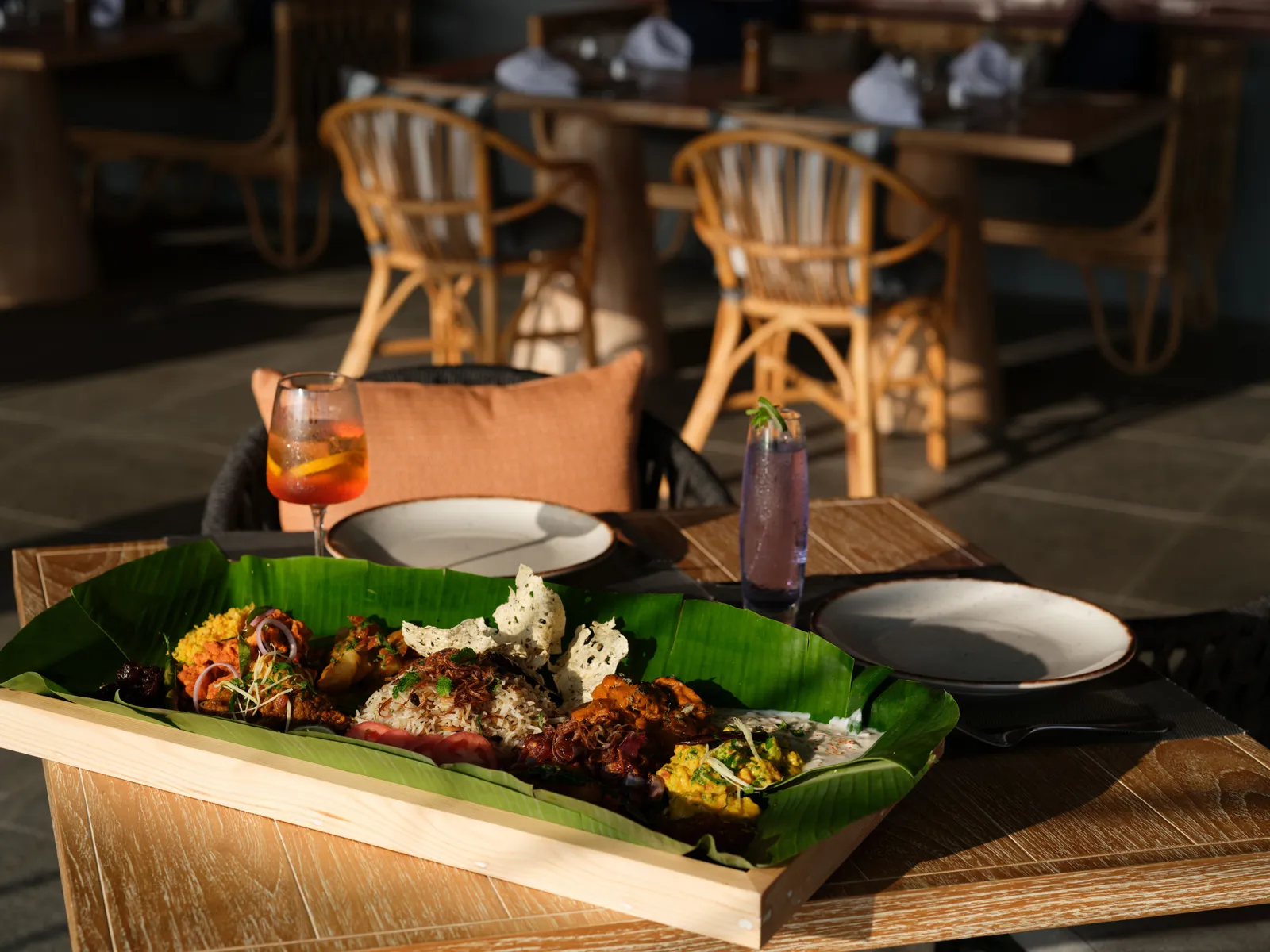 A tray of Sri Lankan dishes on a banana leaf, with 2 cocktails, at a table in Thari.