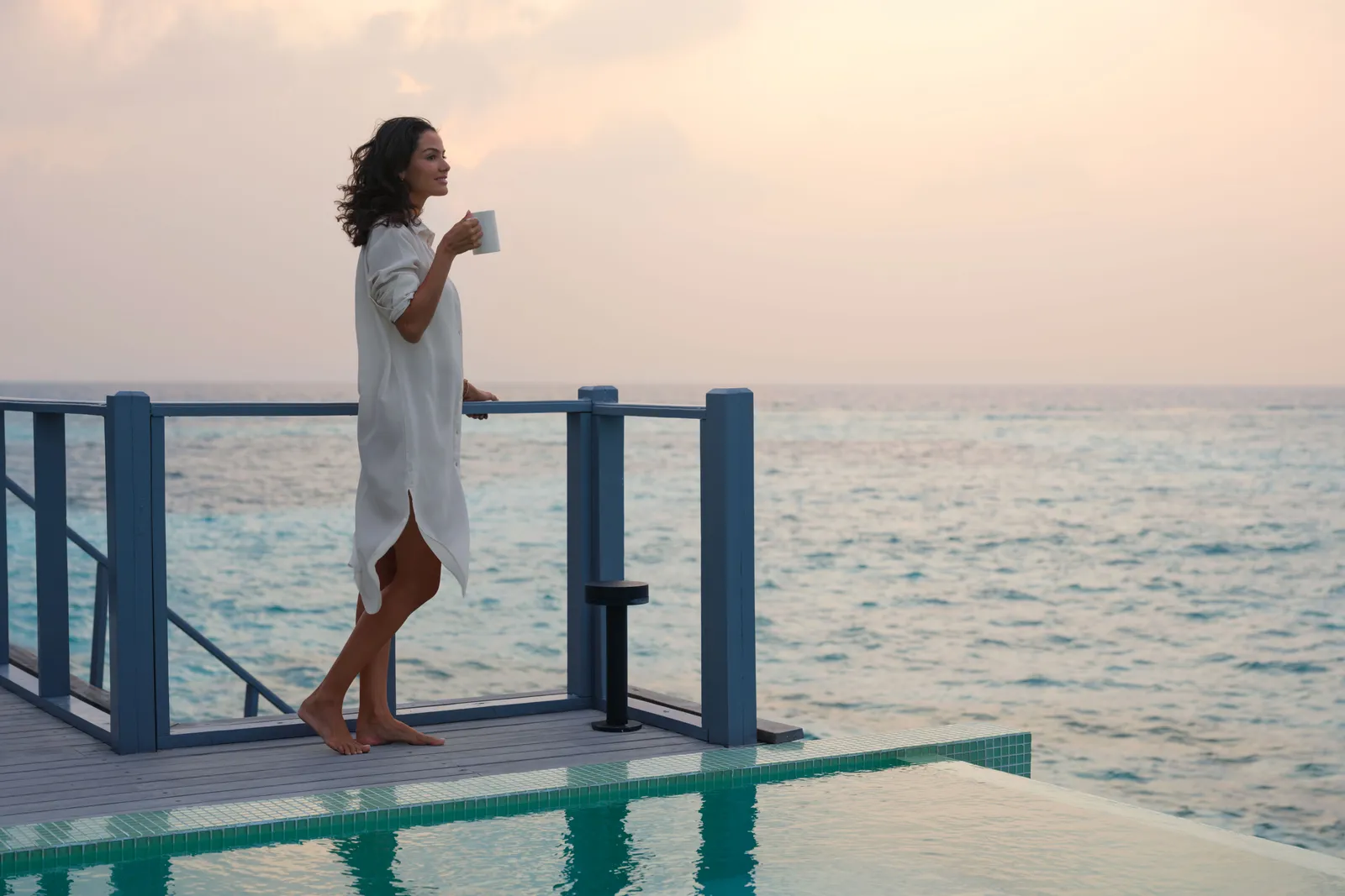 A woman holding a cup on the deck of a water villa at dawn, above the private pool and the sea.