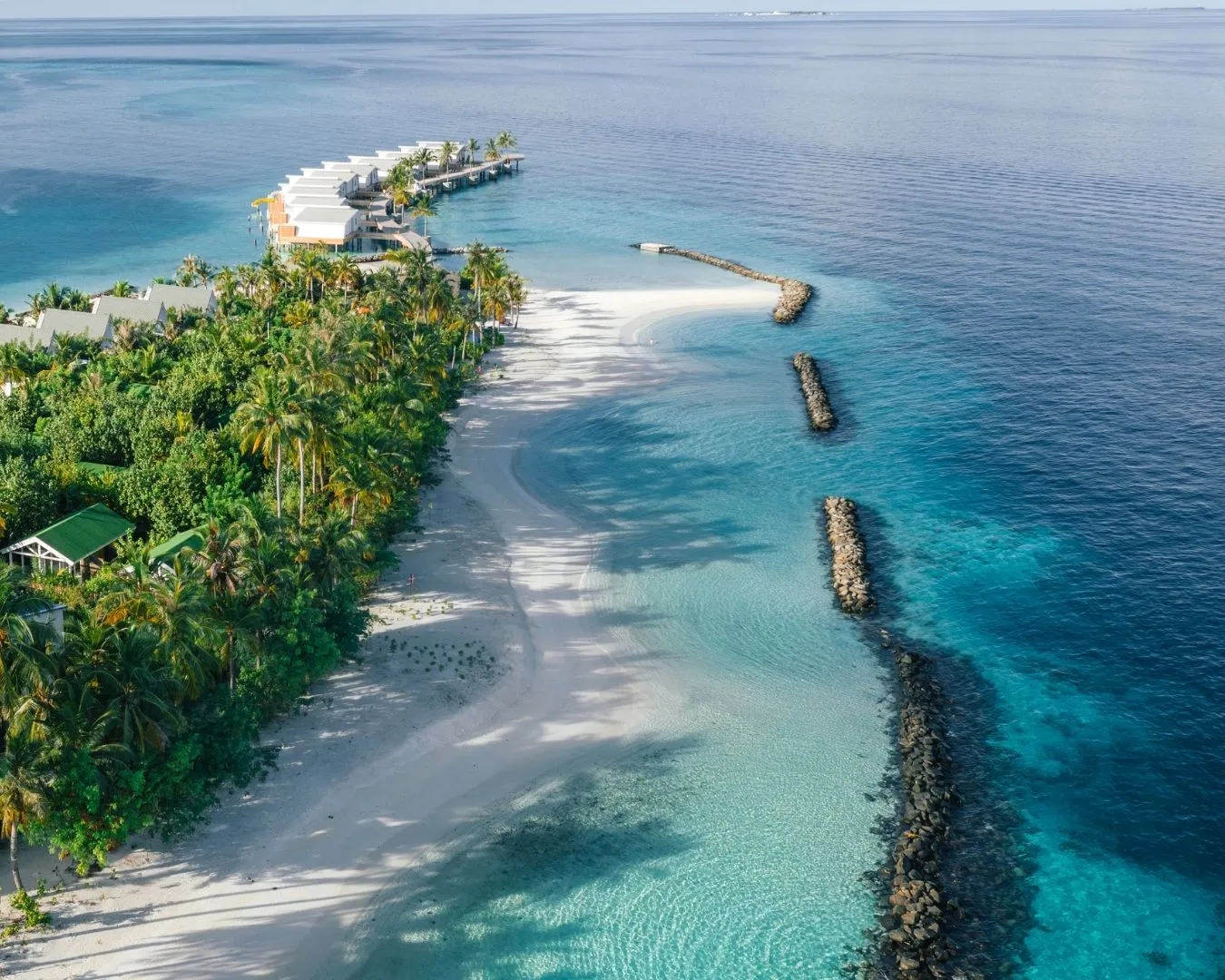 The beach from above: white sand along the shore, the stone breakwaters in the lagoon and the water villas beyond.