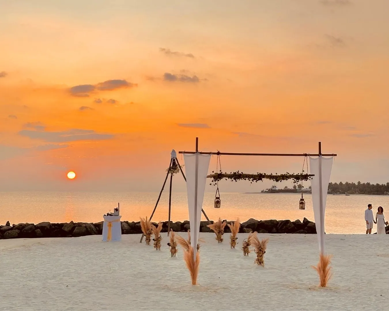 A wooden arch for a beach ceremony at sunset, with lanterns and the low sun over the sea.