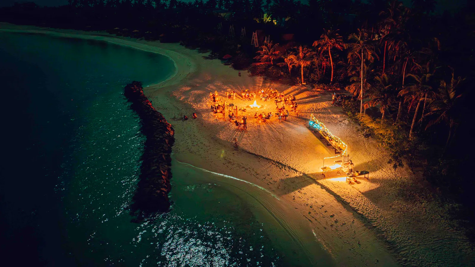 The beach at night from above: a bonfire on the sand, guests around it and the stone breakwater in the dark lagoon.