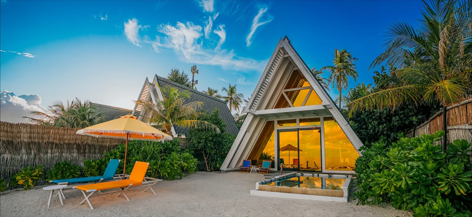 A Bodu Haruge Beach Villa: the A-frame with its lit glass front, the pool before it and loungers under a parasol.