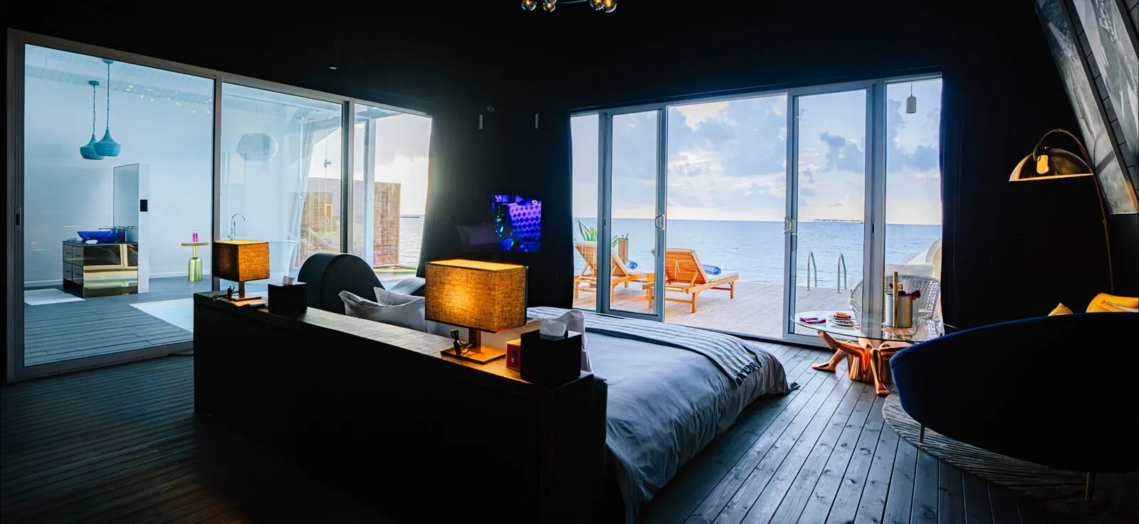 A dark-toned Odi bedroom: the low bed, a blue armchair and glass doors onto the deck and the lagoon.