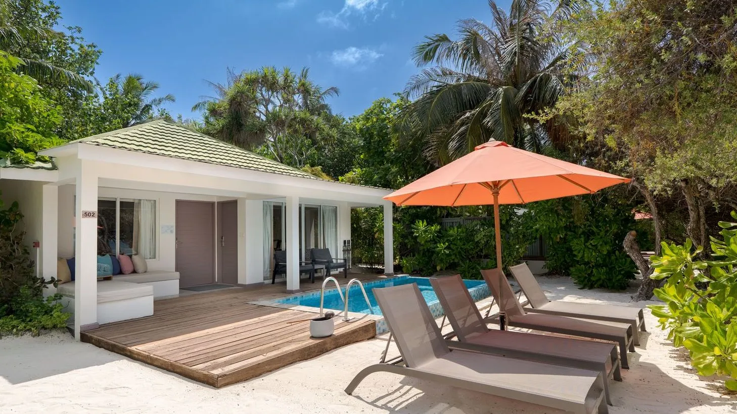 The veranda of a Beach Suite among the palms, with an orange parasol, loungers and the pool