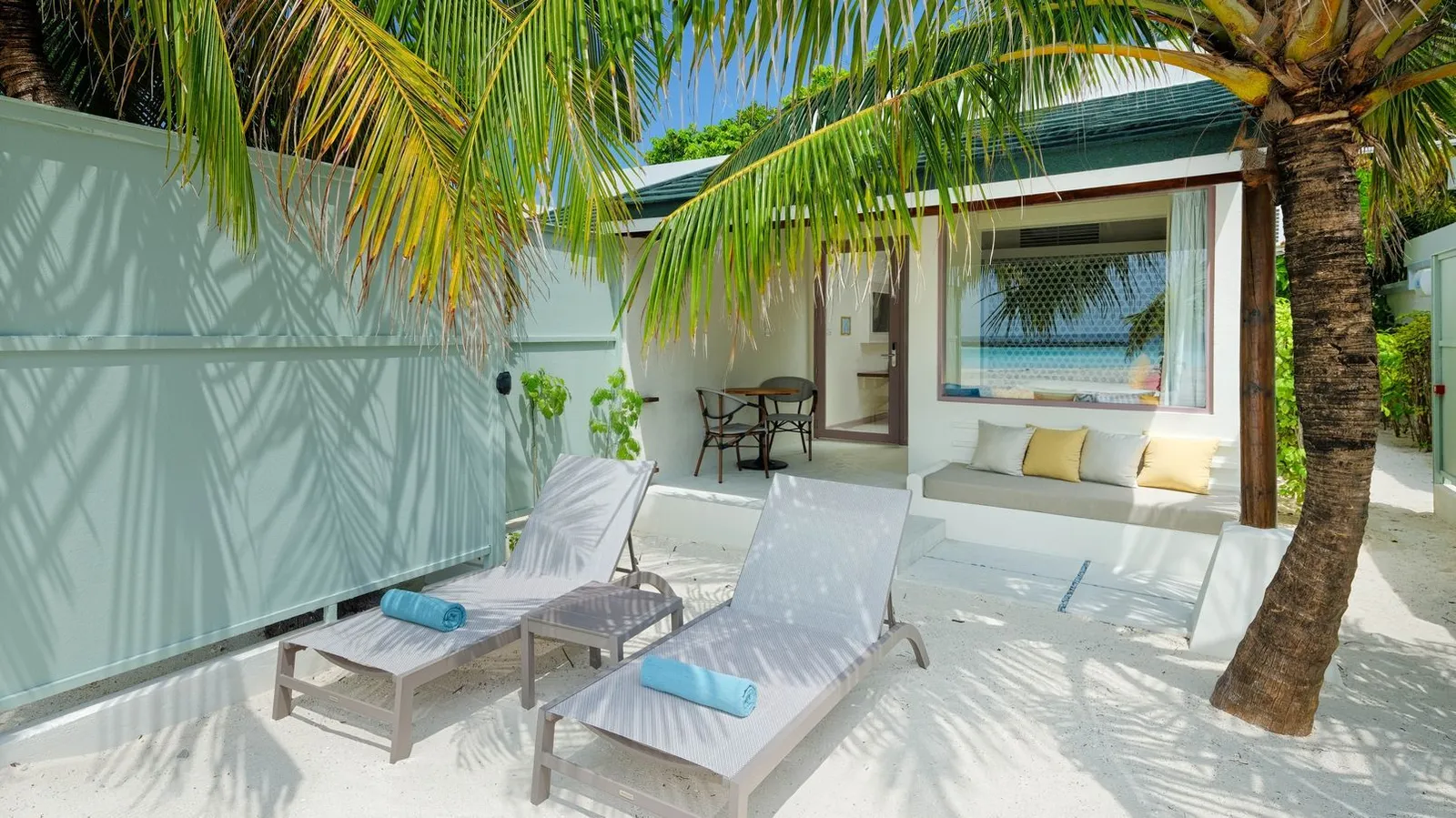 The courtyard of a Deluxe Beach Villa, with 2 loungers under a palm and the veranda; no pool