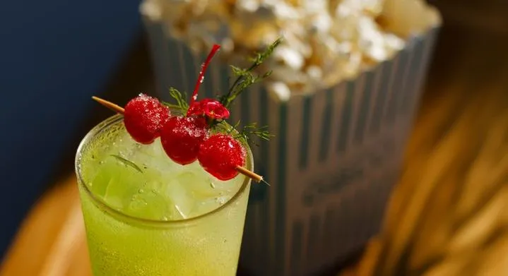 A green cocktail with cherries and a carton of popcorn on a wooden table