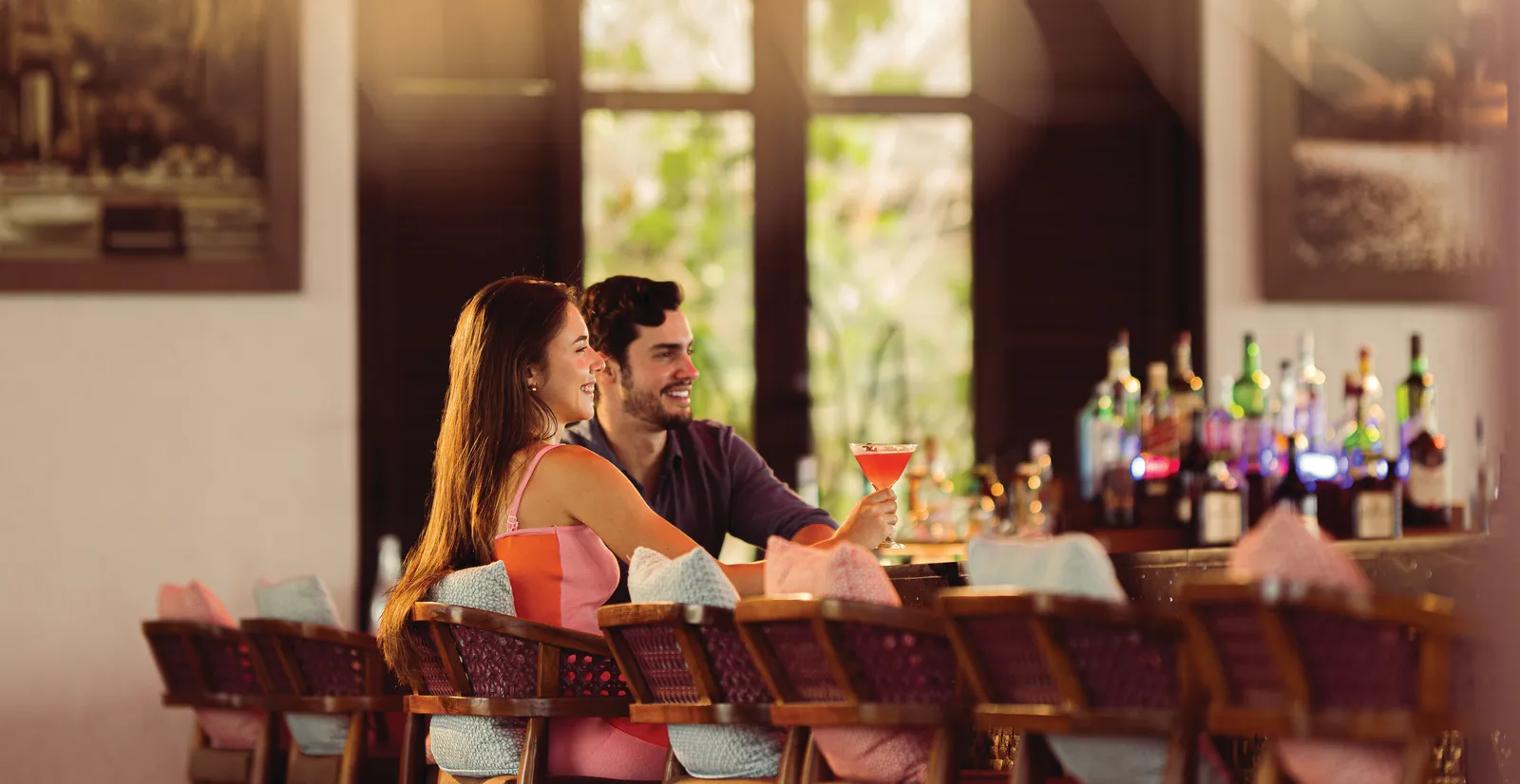 A couple at the bar with 2 cocktails, in front of the shelves of bottles.