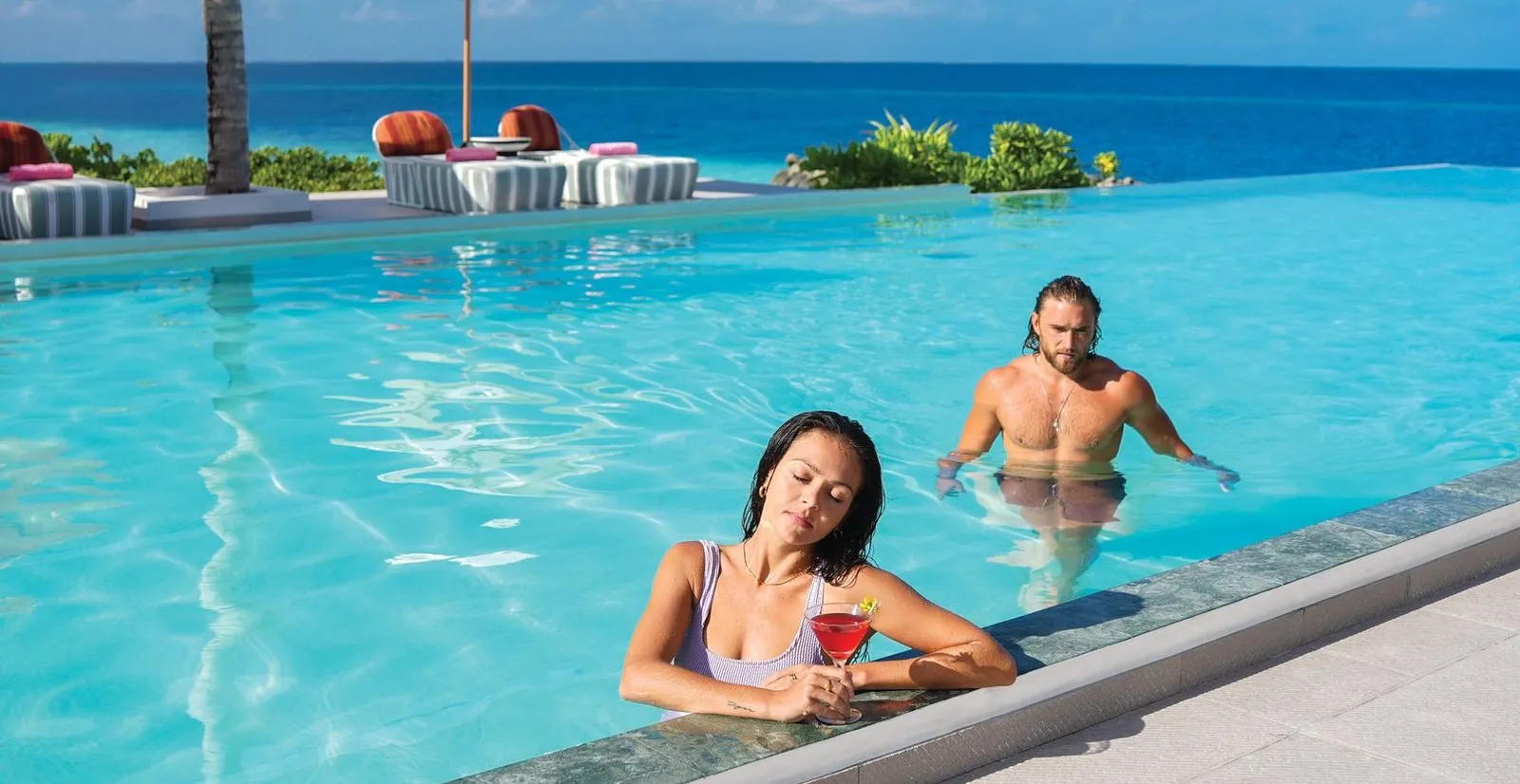A couple in the West Beach Bar’s infinity pool, a cocktail on the edge and the sea beyond the daybeds.