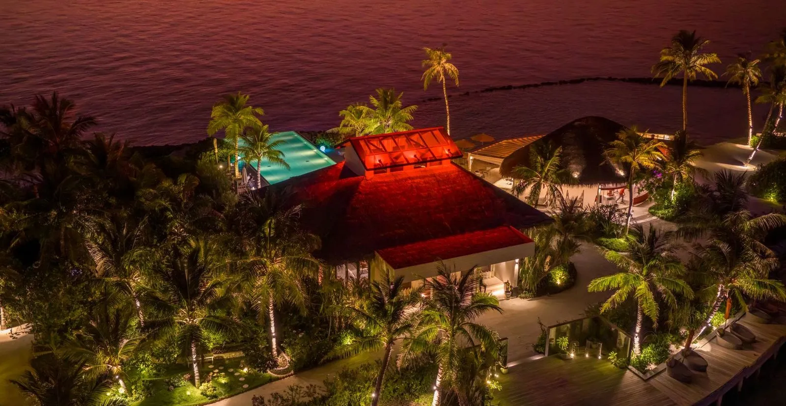 The island from above at night: lit roofs among the palms, the pool and the dark sea.
