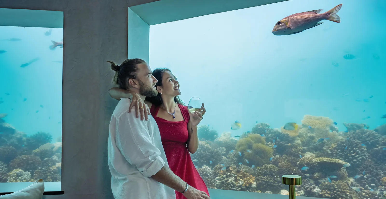 A couple at the window of Only BLU, the restaurant under the sea, with the reef and a passing fish.