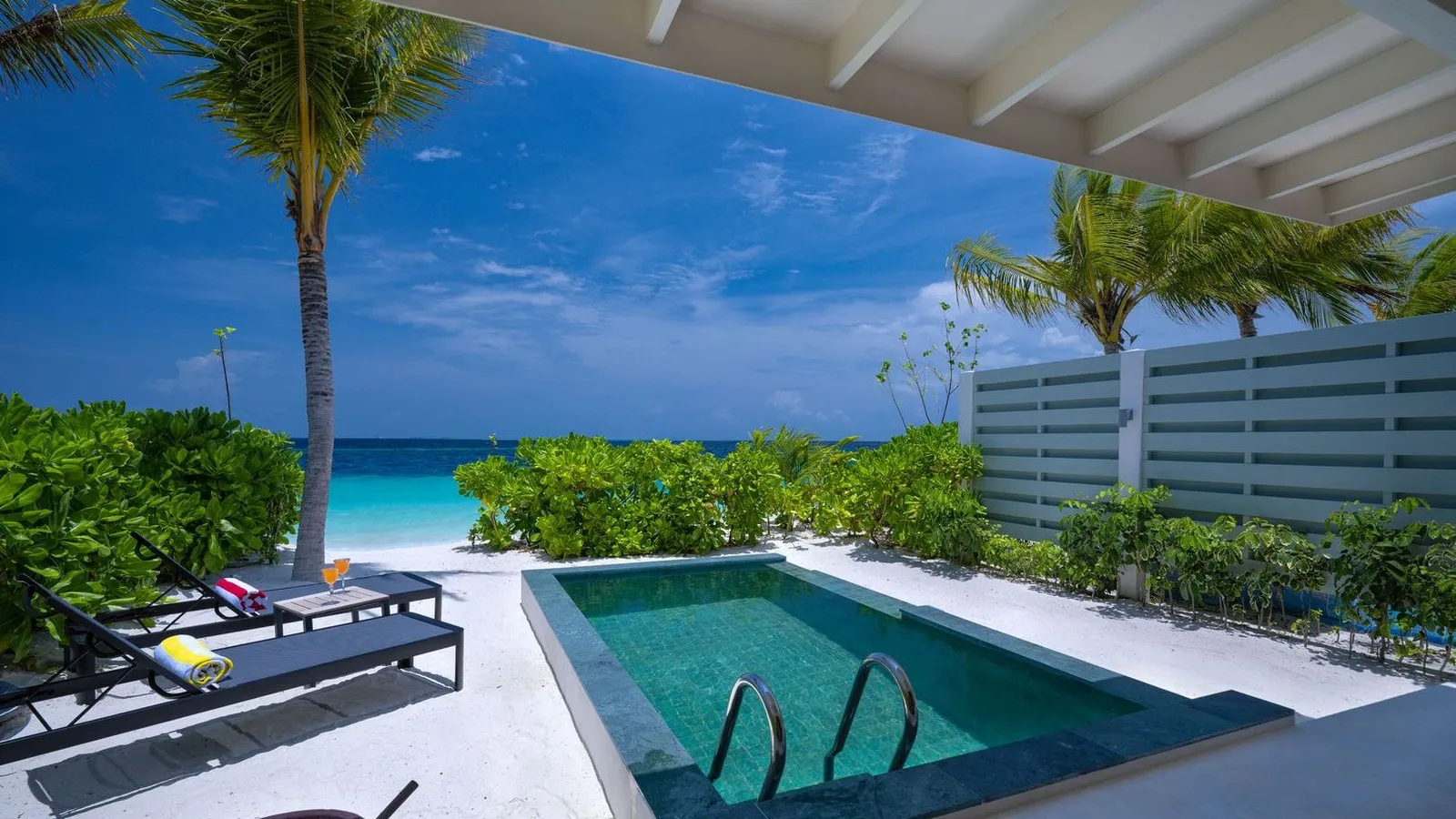 A SunNest Beach Pool Villa’s pool among the palms, with loungers and the beach beyond the hedge.