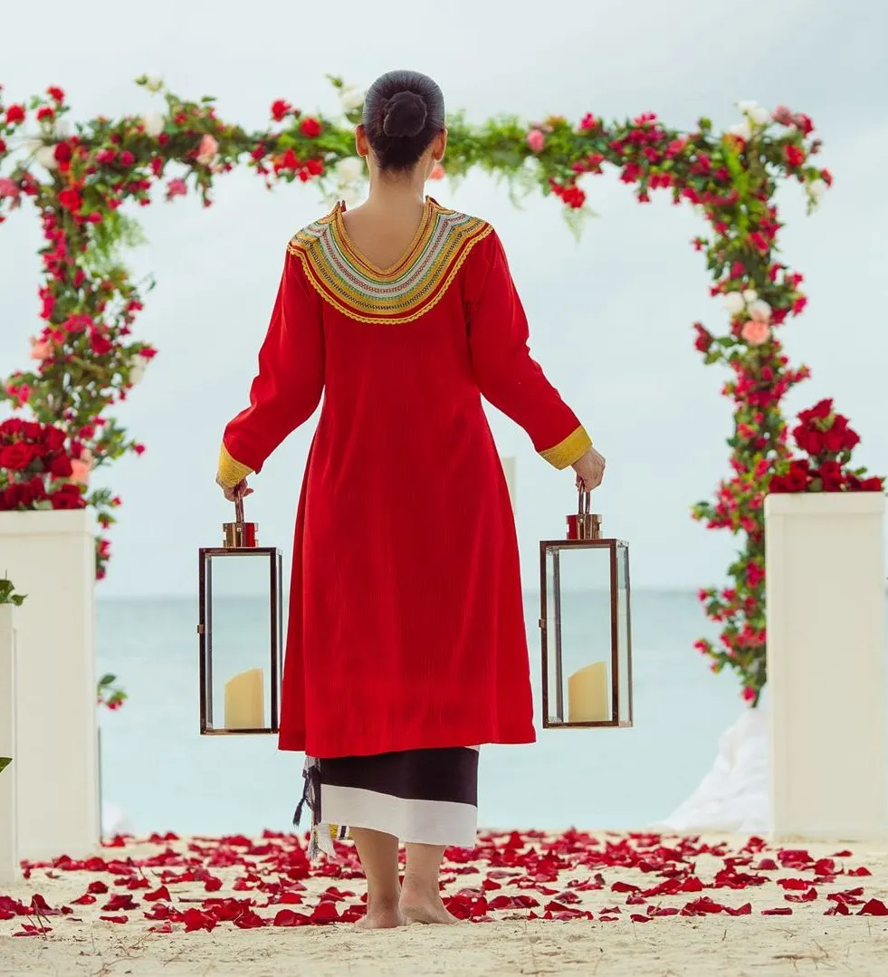 A woman in a red traditional dress, holding 2 lanterns, under the flower arch set on the sand for a vow renewal.