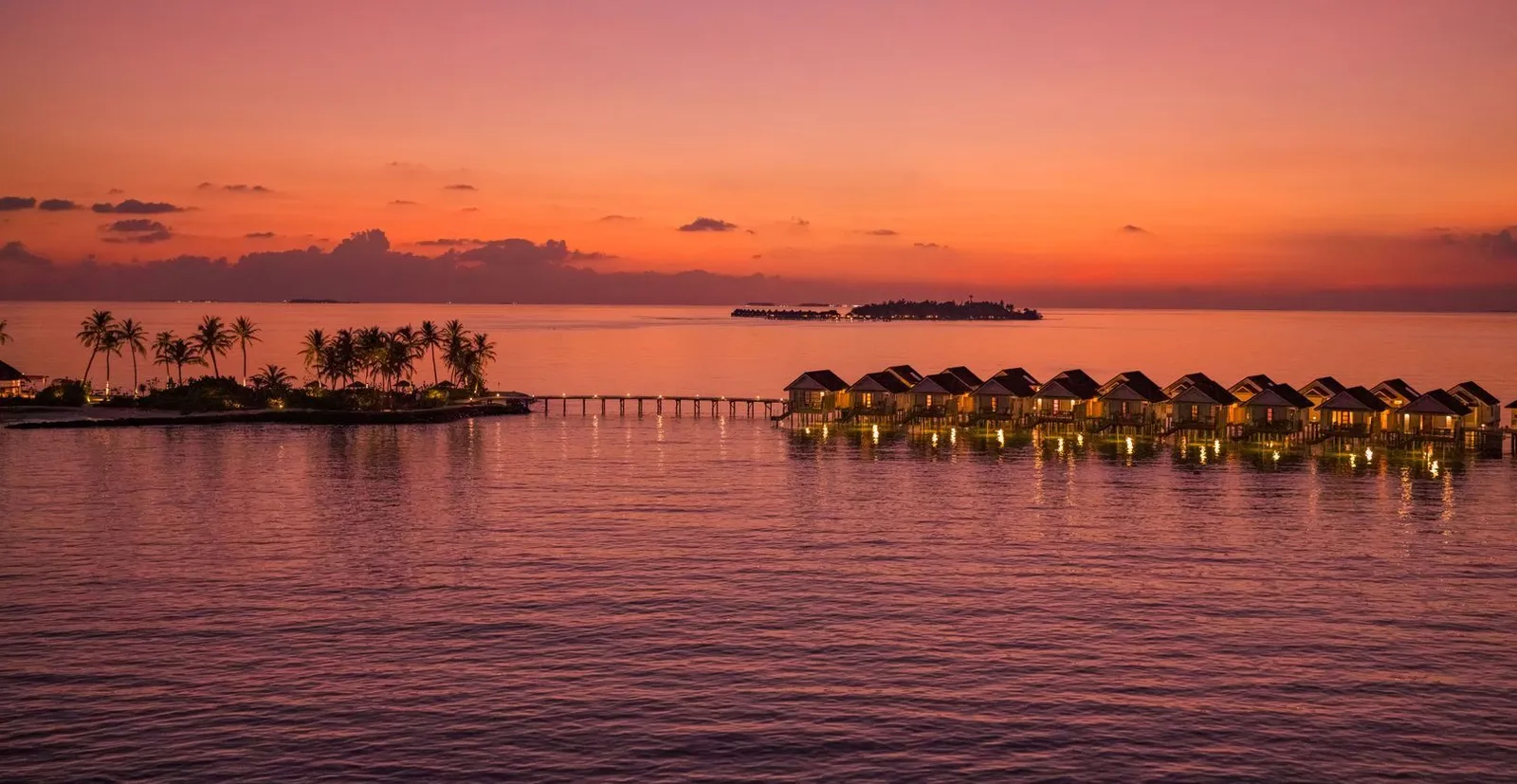 The water villas lit up at sunset, the island’s palms on the left and another island on the horizon.