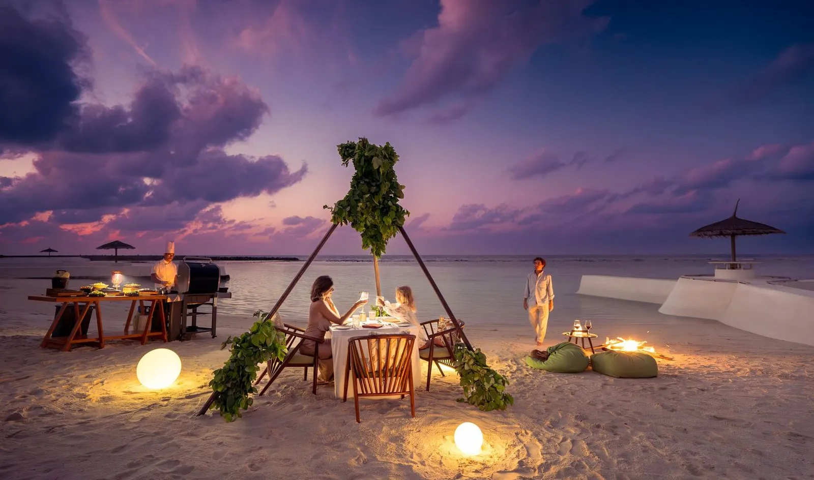 A dinner on the sand at dusk, the table under an arch of leaves, lanterns and a waiter by the water.