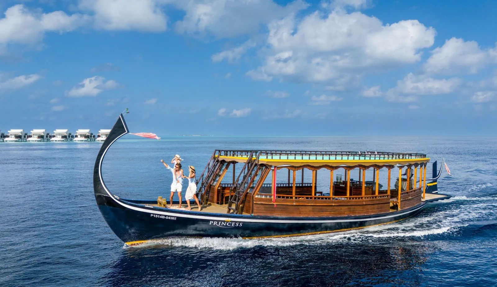 A family on the bow of a traditional dhoni at sea, with the water villas in the distance.