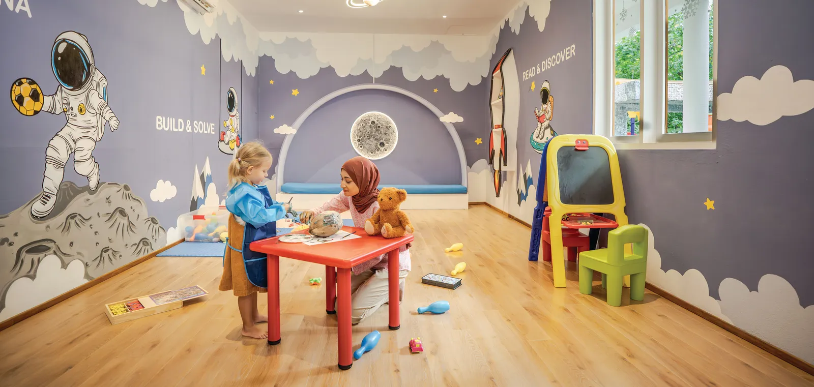 2 children drawing at the kids’ club’s red table, among walls painted with an astronaut and the moon.