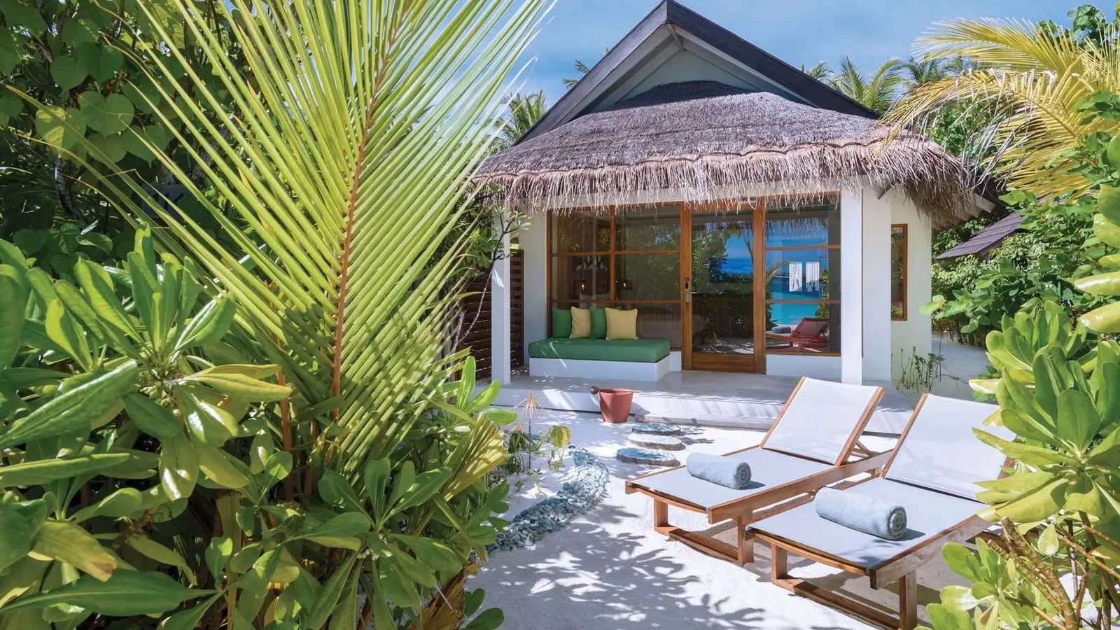 A Beach Villa under its thatched roof among the plants, with the veranda and 2 loungers on the sand.