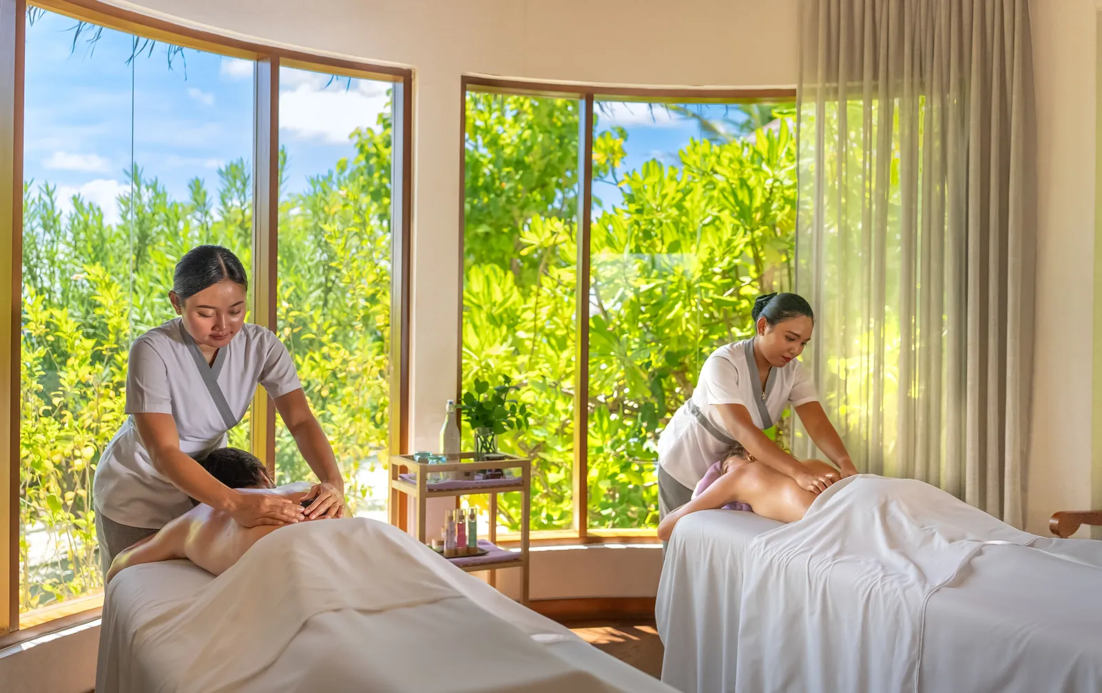 2 ELE|NA Ayur therapists massage a couple in a room with wide windows onto the greenery.