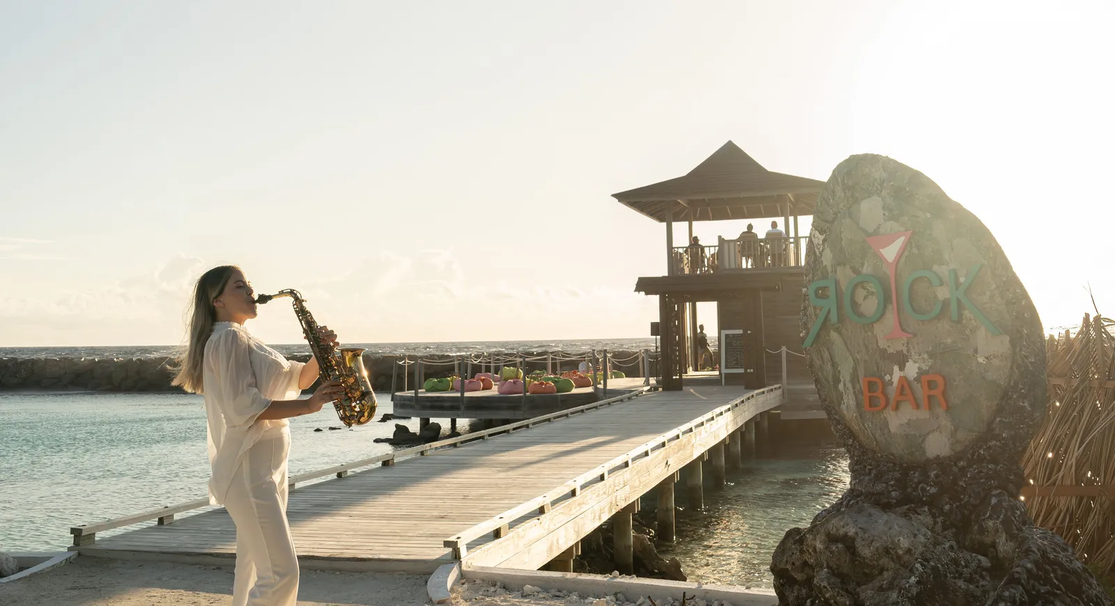 A saxophonist on The Rock’s walkway at sunset, with the bar on stilts and its sign on the rock.