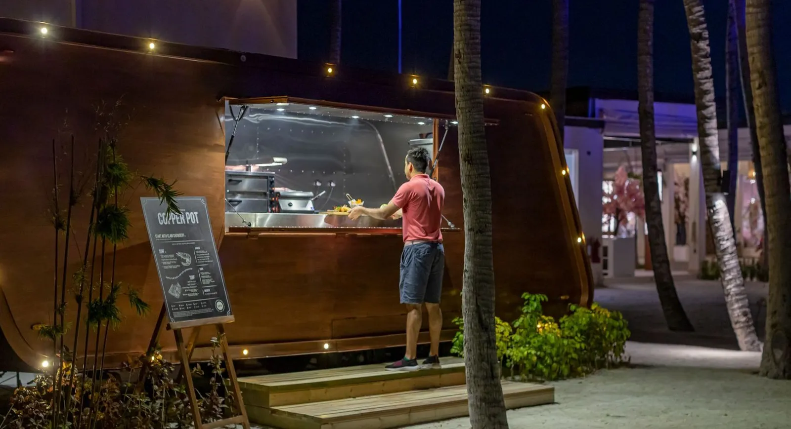 The Copper Pot at night, the food truck among the palms with its open kitchen and menu board.