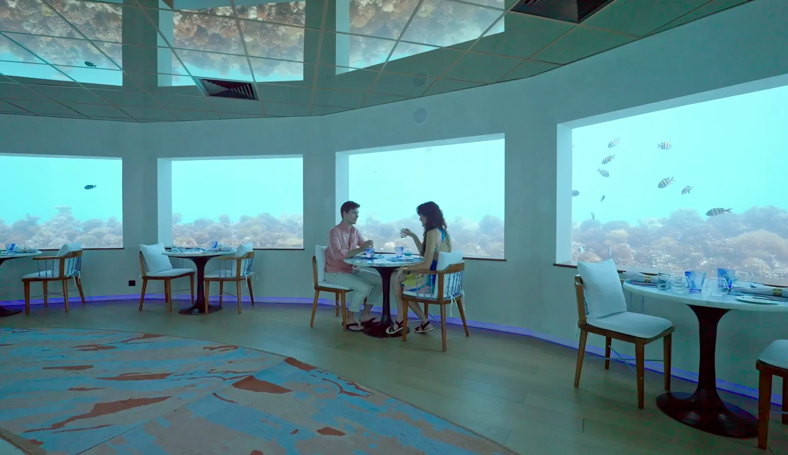 Only BLU’s dining room under the sea: a couple at a table by the windows on the reef, fish drifting past.