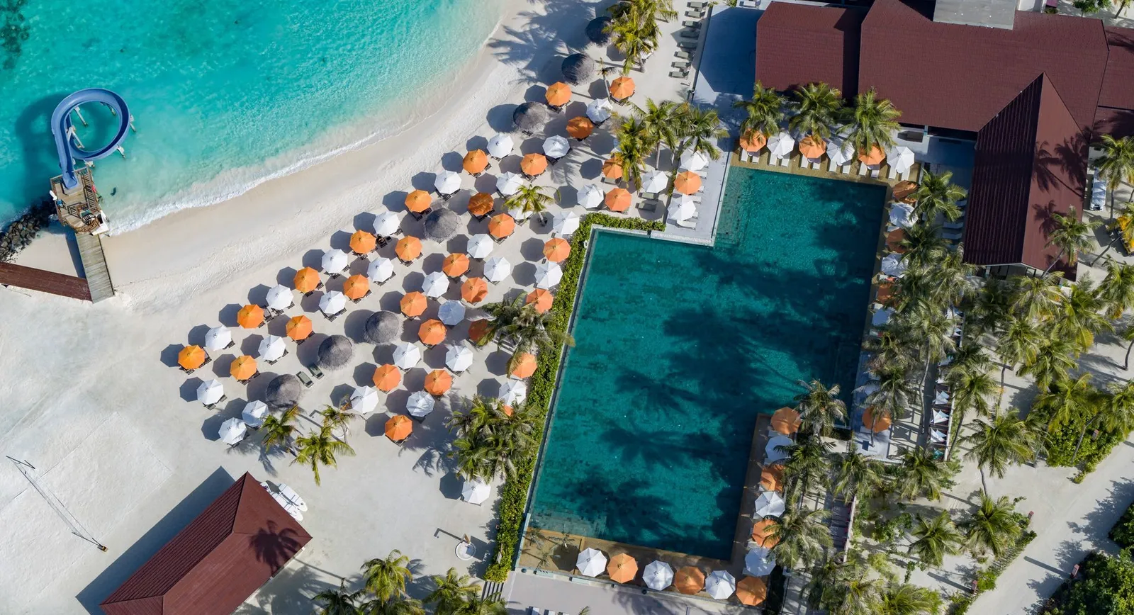 The main pool from above, between the beach with its orange and white parasols and the palms.